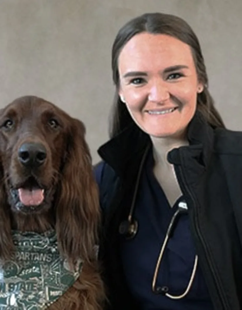 Dr. Katie Haydett Hilton smiling in front of a grey backdrop hugging a large brown dog